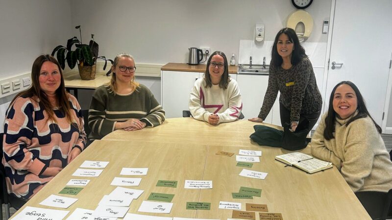 Midwife with a group of attendees at the Healthy Pregnancy Programme. All women are sat around a table smiling.